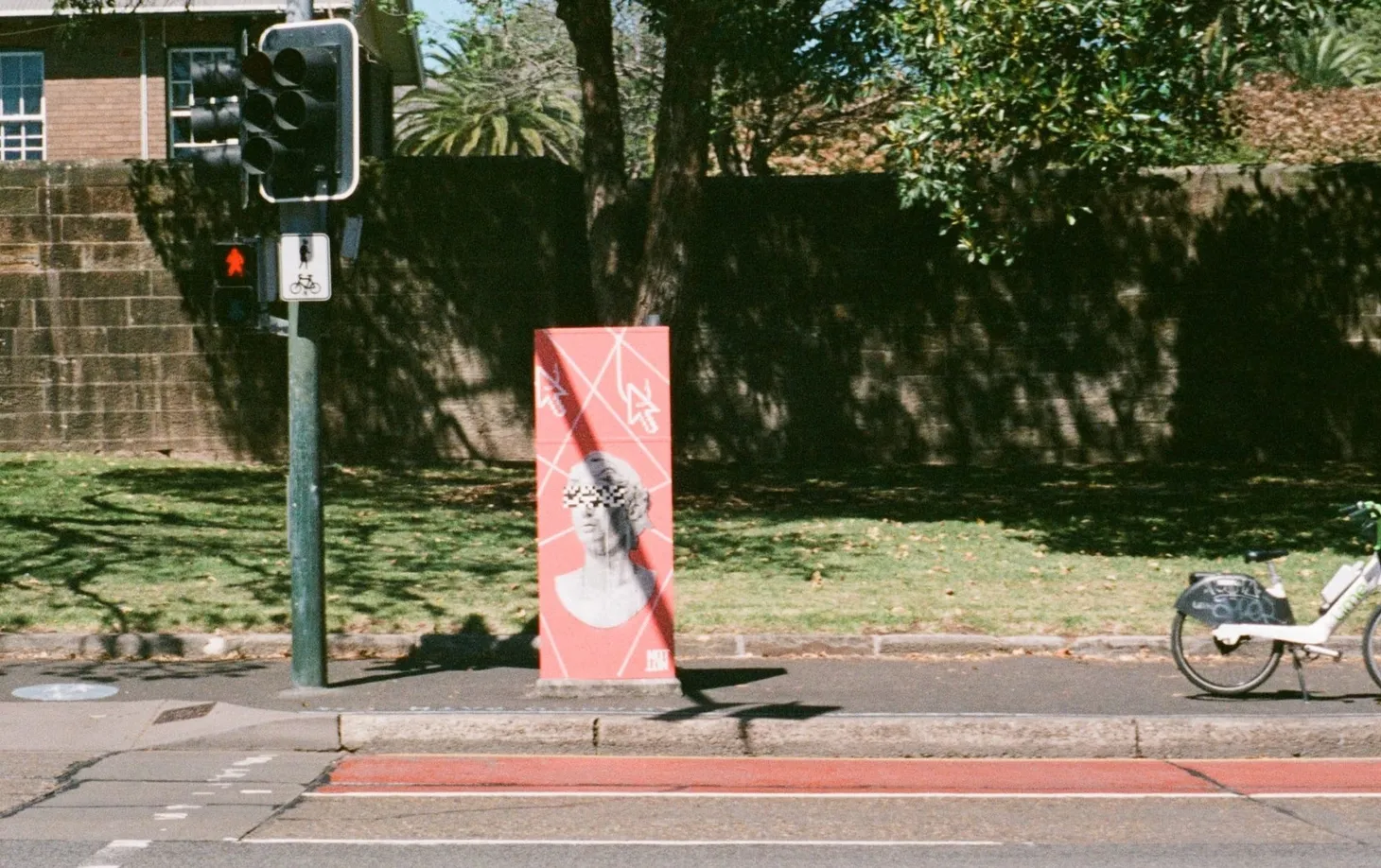 Photo of an electricity box painted pink on the sidewalk featuring a bust with the eyes pixelated next to a red pedestrian do-not-walk sign